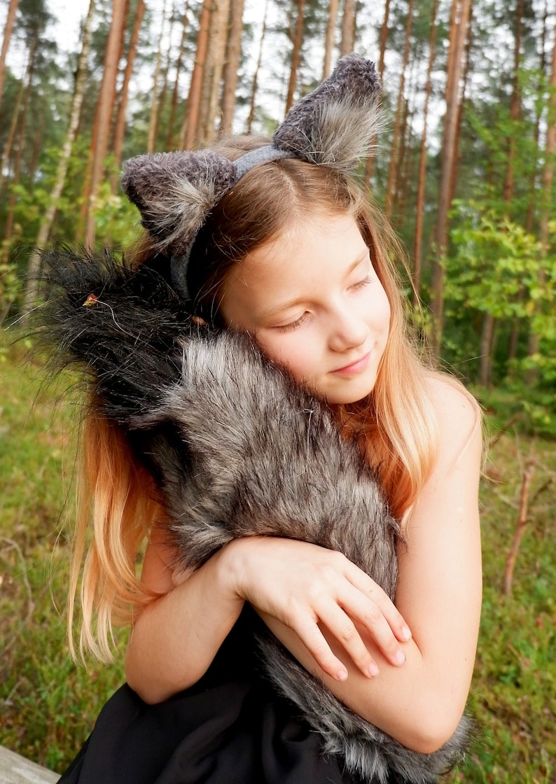 May include: A young person wearing a gray wolf ear headband and holding a gray furry tail prop. The person is in a forest setting.
