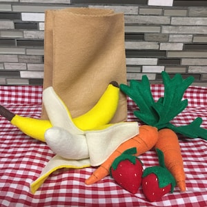 May include: A brown paper bag filled with felt fruit and vegetables. The bag is on a red and white checkered tablecloth. The fruit and vegetables include two bananas, two carrots, and two strawberries.