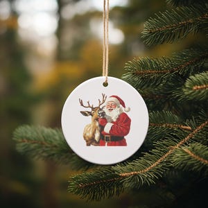May include: A round white Christmas ornament featuring Santa Claus in a red suit and a reindeer. The ornament is hanging from a natural twine string and is set against a blurred background of evergreen branches.