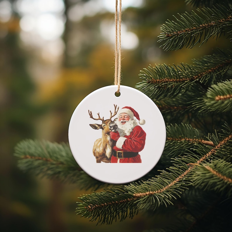 May include: A round white Christmas ornament featuring Santa Claus in a red suit and a reindeer. The ornament is hanging from a natural twine string and is set against a blurred background of evergreen branches.