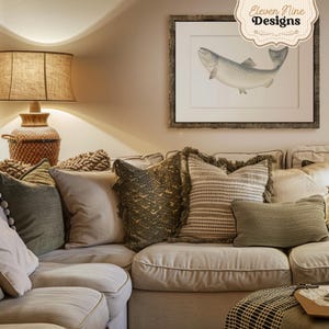 May include: A sectional sofa with a variety of throw pillows in shades of green and beige. The sofa is positioned in front of a wall with a framed print of a fish. The print is framed in a rustic wood frame. The text "Eleven Nine Designs" is visible in the upper right corner of the image.