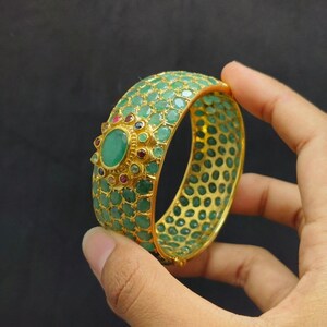 May include: A gold bangle bracelet with a green and red gemstone design. The bracelet is decorated with a large green gemstone in the center.