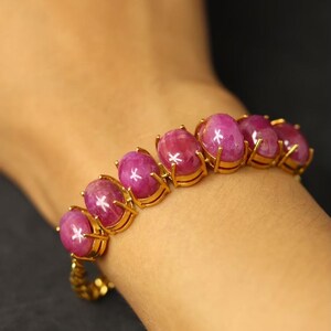 May include: A gold bracelet featuring oval, pink gemstones. The gemstones are set in individual gold settings and are connected by a gold chain. The bracelet is worn on a wrist against a dark background.