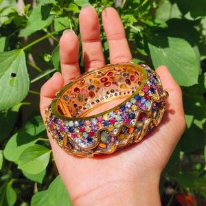 Pode incluir: Uma pulseira dourada cravejada com uma variedade de pedras preciosas coloridas. A pulseira apresenta um design ondulado ao longo das bordas e é mantida contra um fundo de folhagem verde. As pedras preciosas incluem pedras vermelhas, azuis, amarelas e verdes.