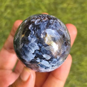 Op de afbeelding: Een gepolijste bolvormige steen met een complex patroon van diep marineblauwe, antracietgrijze en helderwitte wervelingen. Het oppervlak van de bol is glad en reflecterend, met natuurlijke minerale insluitingen en gevarieerde lichtreflecterende texturen.