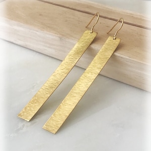 May include: A pair of long, gold-toned earrings with a textured, rectangular design. The earrings are made of metal and have a simple, minimalist style.