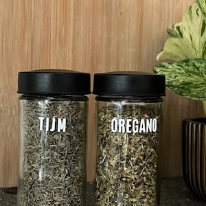 Op de afbeelding: Twee glazen kruidenpotten met zwarte deksels, gevuld met gedroogde kruiden. Eén pot is gelabeld met "TIJM" en de andere met "OREGANO" in witte tekst. De potten staan op een donker gespikkeld oppervlak, met een houten snijplank en een potplant op de achtergrond.