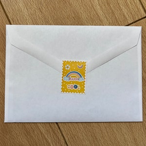 May include: A white envelope with a yellow sticker featuring a rainbow, clouds, and the words "HAPPY COOL". The sticker is in a postage stamp design. The envelope is on a wooden surface.