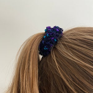 Può includere: Primo piano di un elastico per capelli blu scuro e viola che tiene una coda di cavallo. L'elastico è realizzato in un tessuto strutturato con accenti turchesi. I capelli sono castano chiaro e lisci.