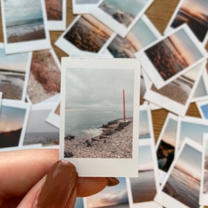 Op de afbeelding: Een verzameling kleine vierkante foto's met kusttaferelen. Eén foto toont een rotsachtige kustlijn met een rode paal, de oceaan en een bewolkte lucht. De andere foto's tonen verschillende strand- en oceaanbeelden.