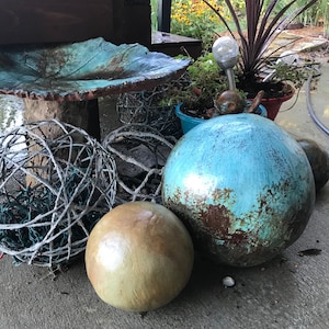 May include: A collection of decorative garden ornaments, including a leaf-shaped birdbath, a large blue sphere, a smaller tan sphere, and a woven sphere. The ornaments are arranged on a concrete patio.