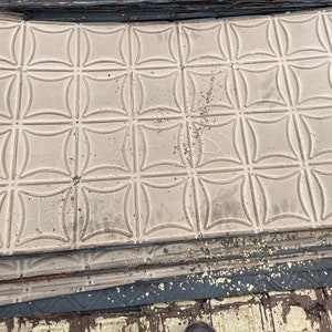 May include: A stack of vintage tin ceiling tiles with a decorative, embossed, quatrefoil pattern. The tiles are a light beige color and have a slightly distressed look.