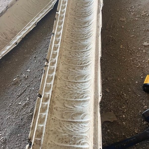 May include: A close-up of a white, decorative, metal ceiling tile with a repeating pattern of swirls. The tile is slightly damaged and shows signs of wear.