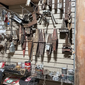 May include: A wall display of various rusty metal hinges and hardware. The hinges are arranged in rows and columns, with some hanging from hooks and others stacked on shelves. The hardware is a mix of different sizes and shapes, and some are painted red. The wall is painted a light beige color.
