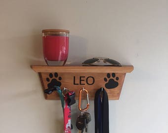 May include: Wooden key holder with paw prints and the name "LEO" engraved. It has a red leash, a black leash, a green carabiner, and keys hanging from it. A red candle and sunglasses are on the shelf above.