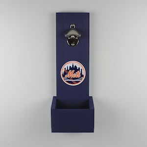 May include: A navy blue wall-mounted bottle opener with a Mets logo. The opener is made of metal and is mounted on a wooden board with a small box at the bottom to catch bottle caps.