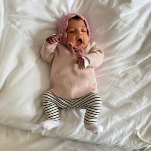May include: A baby wearing a pink knitted hat with a white pom pom, a pink long-sleeved shirt, and black and white striped leggings. The baby is lying on a white sheet.