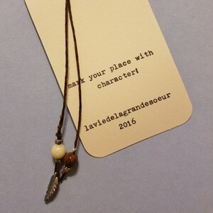 May include: A bookmark with a brown cord and decorative beads. The bookmark has a cream-colored tag with the text "mark your place with character!" and the brand name "laviedelagrandesoeur 2016". A silver feather charm hangs from the cord.