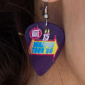 May include: A purple guitar pick earring with a silver hook. The pick features the Vans logo, the number 15, and the text "Warped Tour '09" in yellow and blue. The earring is a unique accessory.