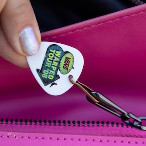 May include: A white guitar pick with the text "Vans Warped Tour '08" in black and green, attached to a black chain. The pick is held against a bright pink background.