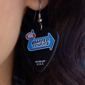 May include: Black guitar pick earring with the text "Vans Warped Tour '05" in white and blue. The earring is made by Dunlop U.S.A. and hangs from a silver fish hook.