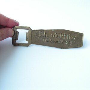 May include: A vintage brass bottle opener with the text "Johnnie Walker Old Scotch Whisky" embossed on the handle. The opener has a rectangular shape with a square opening for removing bottle caps. The brass has a slightly aged appearance.