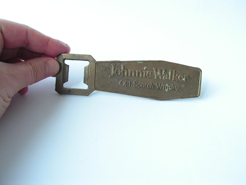 May include: A vintage brass bottle opener with the text "Johnnie Walker Old Scotch Whisky" embossed on the handle. The opener has a rectangular shape with a square opening for removing bottle caps. The brass has a slightly aged appearance.
