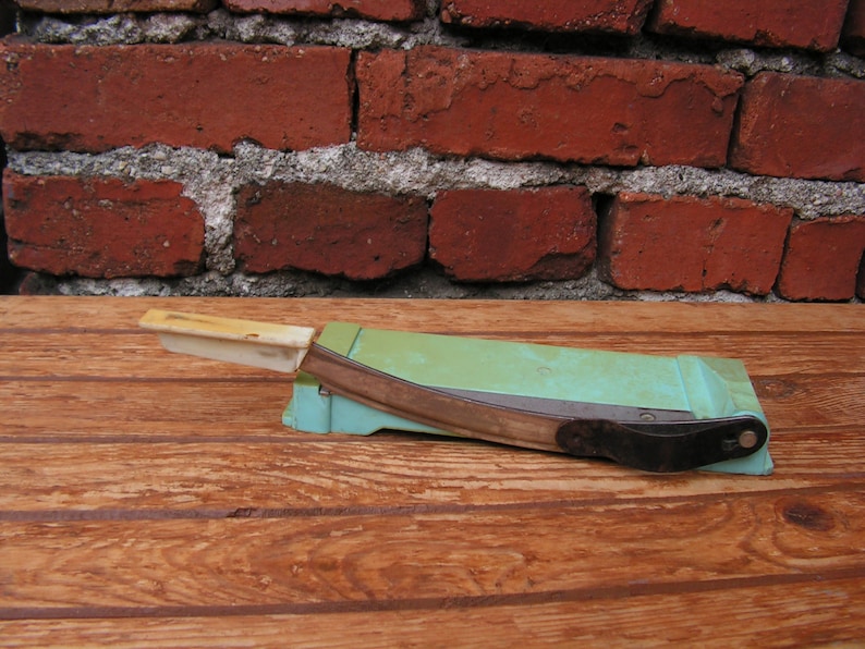 May include: A vintage green and white paper cutter with a metal blade. The cutter is sitting on a wooden surface.