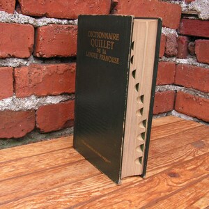 May include: A vintage hardcover dictionary, titled "Dictionnaire Quillet de la Langue Fran&ccedil;aise," stands open on a wooden surface. The book has a dark green cover and a light-colored, notched edge. The background features a weathered red brick wall.
