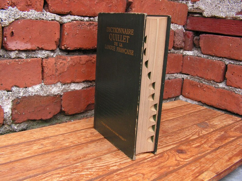 May include: A vintage hardcover dictionary, titled "Dictionnaire Quillet de la Langue Fran&ccedil;aise," stands open on a wooden surface. The book has a dark green cover and a light-colored, notched edge. The background features a weathered red brick wall.