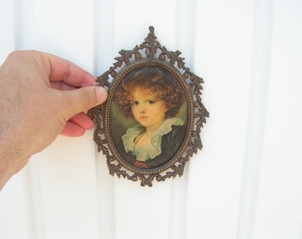 May include: A vintage oval-shaped picture frame with ornate gold metal detailing. The frame holds a portrait of a young girl with curly brown hair wearing a white lace collar.