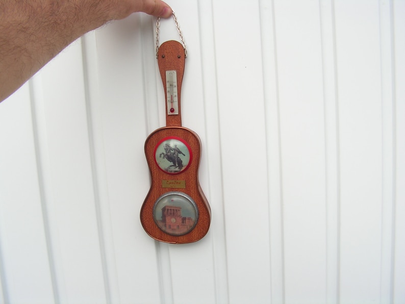 May include: A wooden guitar-shaped thermometer with a red and white thermometer scale, a picture of a cowboy on a horse, and a picture of a building. The thermometer is hanging from a chain.
