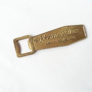 May include: A gold-coloured bottle opener with a rectangular opening and the words "Johnnie Walker Old Scotch Whisky" embossed on the handle.