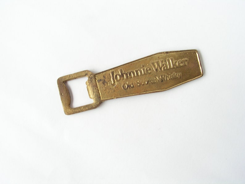 May include: A gold-coloured bottle opener with a rectangular opening and the words "Johnnie Walker Old Scotch Whisky" embossed on the handle.