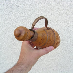 K&ouml;nnte beinhalten: Eine Hand h&auml;lt eine braune Holzflasche mit einem geschnitzten Design und einem Lederriemen.