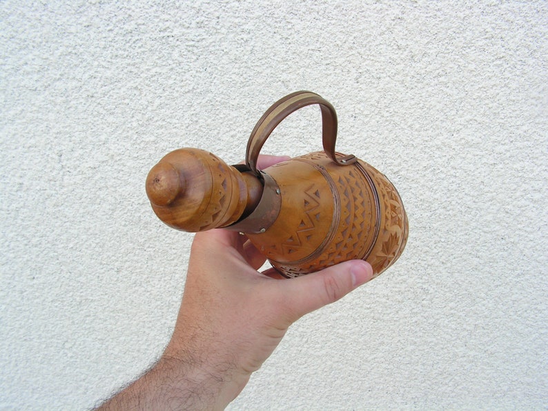 K&ouml;nnte beinhalten: Eine Hand h&auml;lt eine braune Holzflasche mit einem geschnitzten Design und einem Lederriemen.