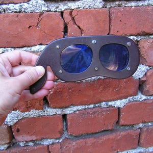 May include: A pair of brown leather goggles with blue tinted lenses. The goggles are held in front of a brick wall.
