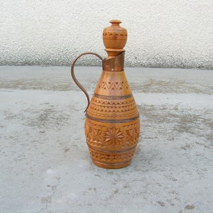 K&ouml;nnte beinhalten: Eine Holzflasche mit einem geschnitzten Design und einem kupferfarbenen Metallgriff. Die Flasche hat eine runde, bauchige Form und einen schmalen Hals. Die Flasche steht auf einer grauen Betonfl&auml;che.
