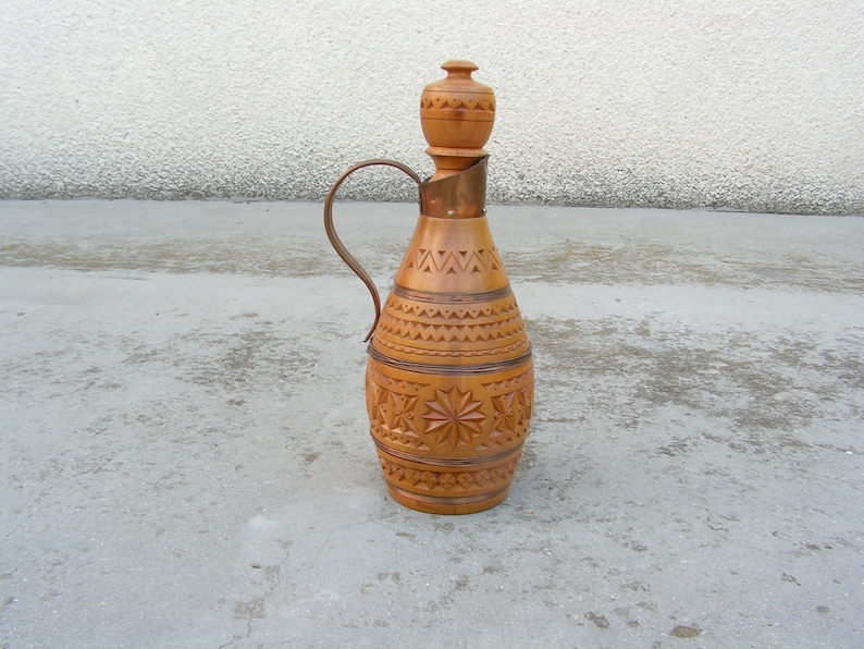 K&ouml;nnte beinhalten: Eine Holzflasche mit einem geschnitzten Design und einem kupferfarbenen Metallgriff. Die Flasche hat eine runde, bauchige Form und einen schmalen Hals. Die Flasche steht auf einer grauen Betonfl&auml;che.