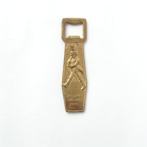 May include: A gold-coloured metal bottle opener with a raised image of a man walking. The opener has the text "Johnnie Walker" and "Scotch Whisky" on it.