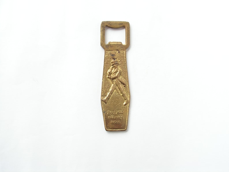 May include: A gold-coloured metal bottle opener with a raised image of a man walking. The opener has the text "Johnnie Walker" and "Scotch Whisky" on it.