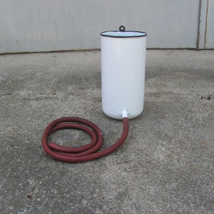 May include: White enamelware pitcher with a black rim and a red rubber hose attached to the spout. The pitcher is sitting on a gray concrete surface.