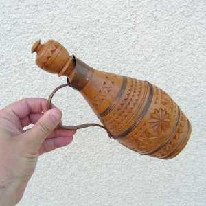 K&ouml;nnte beinhalten: Eine Holzflasche mit einem geschnitzten Design und einem Metallgriff. Die Flasche ist braun und hat einen hellbraunen Holzstopfen.