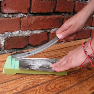 May include: A green and white plastic paper trimmer with a silver blade. The trimmer is on a wooden surface and a person is using it to cut a black and white photo.