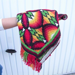 May include: A colorful, embroidered textile with a geometric floral pattern in red, orange, yellow, and green. The edges are trimmed with a red fringe. The textile is held up, showcasing its intricate design and craftsmanship.