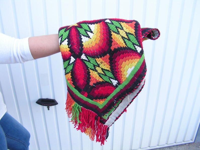 May include: A colorful, embroidered textile with a geometric floral pattern in red, orange, yellow, and green. The edges are trimmed with a red fringe. The textile is held up, showcasing its intricate design and craftsmanship.