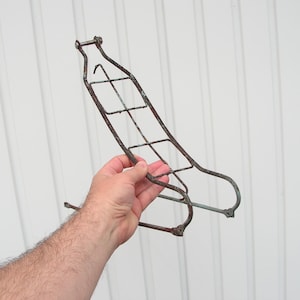 May include: A rusty, vintage bicycle rack, held by a hand. The rack is made of metal and has a grid-like design with curved edges. It appears to be an older model, showing signs of age and wear, with a faded brown colour.