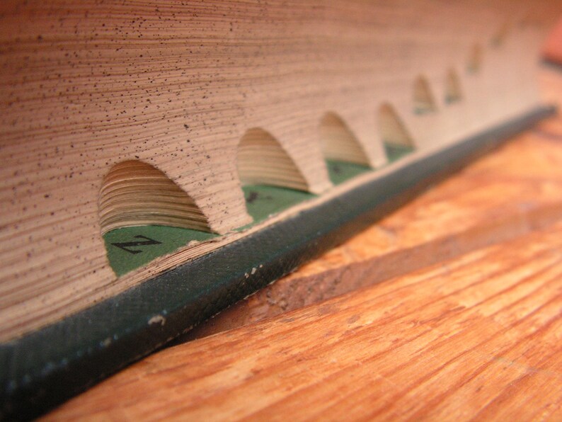May include: Close-up of a vintage book with arched cutouts along the edge. The pages are aged with a speckled texture, and green markers are visible within the cutouts. The book's spine is dark green, resting on a wooden surface.