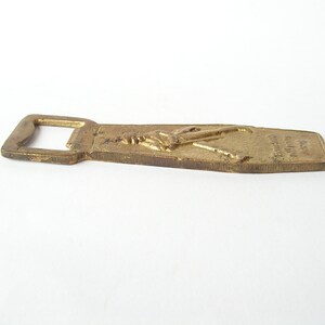 May include: A brass bottle opener featuring a raised design of a person holding a bottle of beer. The opener is engraved with the words "Budweiser" and "King of Beers".