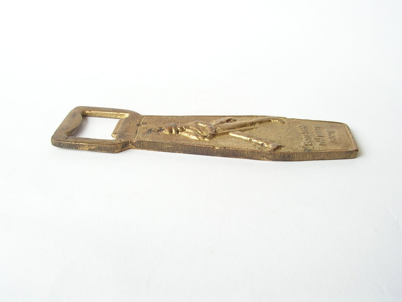 May include: A brass bottle opener featuring a raised design of a person holding a bottle of beer. The opener is engraved with the words "Budweiser" and "King of Beers".